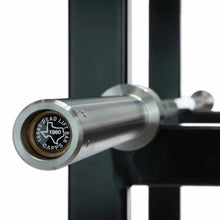 Load image into Gallery viewer, Close-up of a stainless steel barbell end with a circular black and gold medallion engraved with 'TEXAS DEAD LIFT BAR CAPPS 1980' and the state of Texas outline, mounted on a black weightlifting rack.